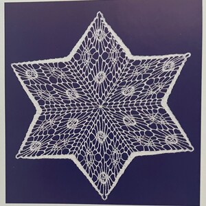 May include: A white, star-shaped doily with a delicate, openwork design. The intricate pattern features a central point with radiating lines and circular accents. The doily is set against a dark blue background, highlighting its detailed craftsmanship.