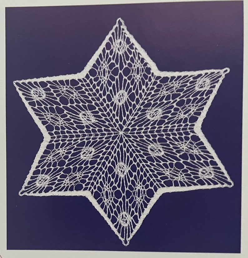 May include: A white, star-shaped doily with a delicate, openwork design. The intricate pattern features a central point with radiating lines and circular accents. The doily is set against a dark blue background, highlighting its detailed craftsmanship.