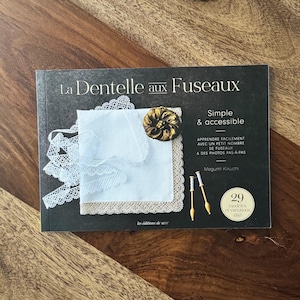 May include: A book titled "La Dentelle aux Fuseaux" with a dark grey cover, featuring a white lace handkerchief, a gold-coloured button, and text. The book is on a wooden surface. The book's title translates to "Bobbin Lace".