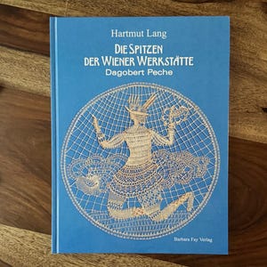 Peut inclure: Livre à couverture rigide bleue intitulé "Die Spitzen Der Wiener Werkstätte Dagobert Peche" de Hartmut Lang. Il présente un motif de dentelle circulaire et du texte en allemand. Posé sur une surface en bois.