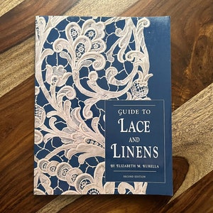 Peut inclure: Un livre bleu avec un motif de dentelle blanche sur la couverture. Le titre est "Guide to Lace and Linens" par Elizabeth M. Kurella. Le livre est une deuxième édition.