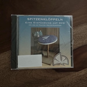 Peut inclure: Un boîtier de DVD avec le titre "SPITZENKLÖPPELN" et le sous-titre "Eine Einführung auf DVD" est présenté. Le boîtier présente une image d'un petit tabouret avec un coussin bleu et un motif de ventilateur décoratif. Le boîtier est dans une pochette en plastique transparent.