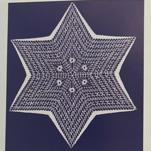 May include: A white, six-pointed star doily with a delicate, openwork design. The star is centered on a dark blue background, creating a striking contrast. The intricate lace-like pattern is visible throughout the star's form.