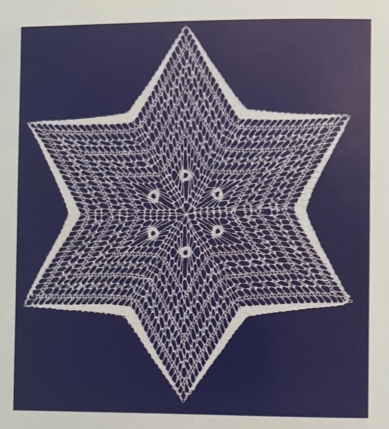 May include: A white, six-pointed star doily with a delicate, openwork design. The star is centered on a dark blue background, creating a striking contrast. The intricate lace-like pattern is visible throughout the star's form.
