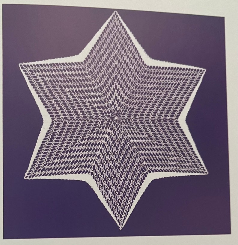 May include: A white, crocheted star doily with a six-pointed design. The intricate openwork pattern is set against a dark blue background. The doily's edges are clearly defined, showcasing the detailed craftsmanship.
