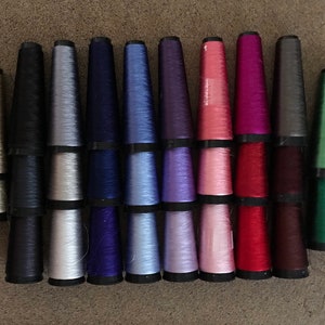 May include: A collection of 16 spools of thread in various colors, including beige, black, silver, blue, purple, pink, red, maroon, and green.