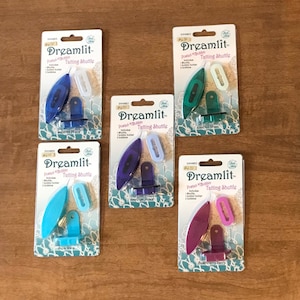 May include: Five different colored Dreamlit Posted-Bobbin Tatting Shuttles. The shuttles are packaged in clear plastic with a white background and black text. The colors are: Amethyst Pearl, Emerald Green, Turquoise Wave, Amethyst Pearl, and Blackberry Berry.