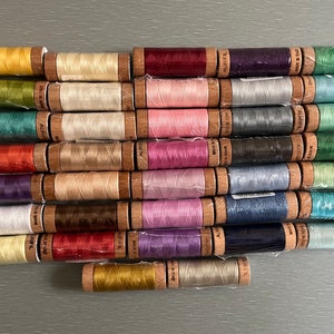May include: A collection of 24 spools of thread in various colors, including yellow, green, red, pink, purple, blue, and gray. The spools are arranged in rows and are all wrapped in wooden bobbins.