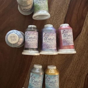 May include: Eight spools of Liz Metallic thread by Handy Hands in various colors including silver, purple, pink, grey, and gold. The labels indicate sizes 10, 20, and 40, noting the thread is strong and kink resistant for crochet and tatting.