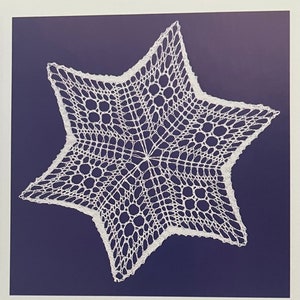 May include: A white, star-shaped doily with intricate lace detailing. The doily is set against a dark blue background, creating a contrast that highlights the delicate openwork pattern. The star has five points and a detailed, open weave design.