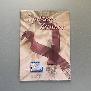 May include: A book titled "Spitzen-Zauber" with a cover featuring a flowing burgundy ribbon and a lace design. The book is wrapped in clear plastic. The cover also has a small sticker with handwritten text.