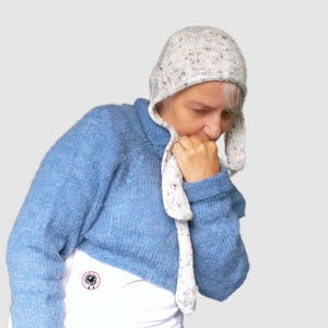 May include: A light blue knitted sweater with long sleeves and a high collar, paired with a speckled off-white bonnet featuring long side ties. The sweater has a small circular logo on the lower front depicting a black sheep illustration.