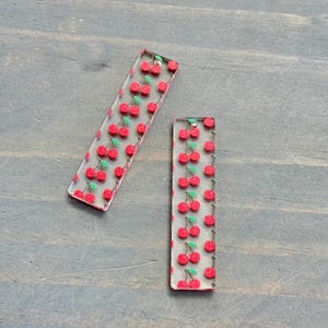 May include: Two clear acrylic rectangular keychains with a red cherry print.