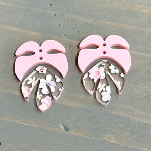 May include: A pair of pink and floral patterned acrylic earrings. The earrings are shaped like a leaf with a pink overlay.