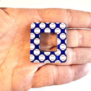 May include: A square-shaped acrylic charm with a blue background and a white outline. The charm is decorated with a repeating pattern of white baseballs with red stitching. The charm has a square cutout in the center.