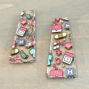May include: Two clear, trapezoid-shaped earrings with a medical theme. The earrings feature a repeating pattern of medical symbols, including a stethoscope, pills, a heart, a medical kit, and a face mask. The colors are pink, blue, and white.