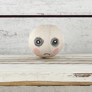 May include: A wooden doll head with a painted face. The doll has large, black eyes, a small, sad mouth, and rosy cheeks.