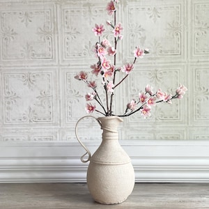 Op de afbeelding: Een crèmekleurige kan met een handvat bevat takken met roze bloemen. De vaas heeft een textuur en staat op een houten oppervlak. De achtergrond is een witte, patroonmuur.