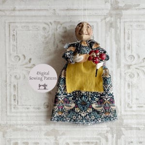 Peut inclure: Poupée en tissu faite à la main, au design fantaisiste. La poupée porte une robe à motifs avec un tablier jaune et tient un bouquet de champignons rouges. Le visage de la poupée a un aspect vieilli unique. Le texte "Digital Sewing Pattern" est inclus.