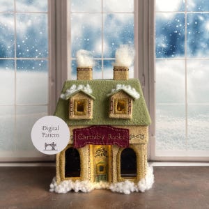 Puede incluir: Una casa de fieltro hecha a mano con un tejado verde, dos chimeneas con humo y un letrero que dice "Carnaby Books". La casa tiene ventanas iluminadas y está decorada con nieve artificial. Hay un patrón digital disponible.