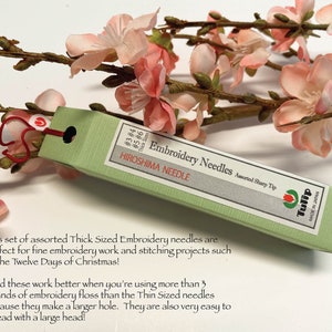 Puede incluir: Una caja verde de agujas de bordado Tulip etiquetada como "Thick Sized" y "Assorted Sharp Tip". La caja est&aacute; hecha en Jap&oacute;n y tiene el texto "Hiroshima Needle" y "Embroidery Needles".