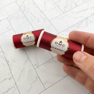 May include: Two spools of DMC Diamant thread in a deep red color. The spools have white cores and gold and white labels. The thread is held by a hand, with one spool in focus and the other slightly blurred in the background. The background is a white and gray patterned surface.