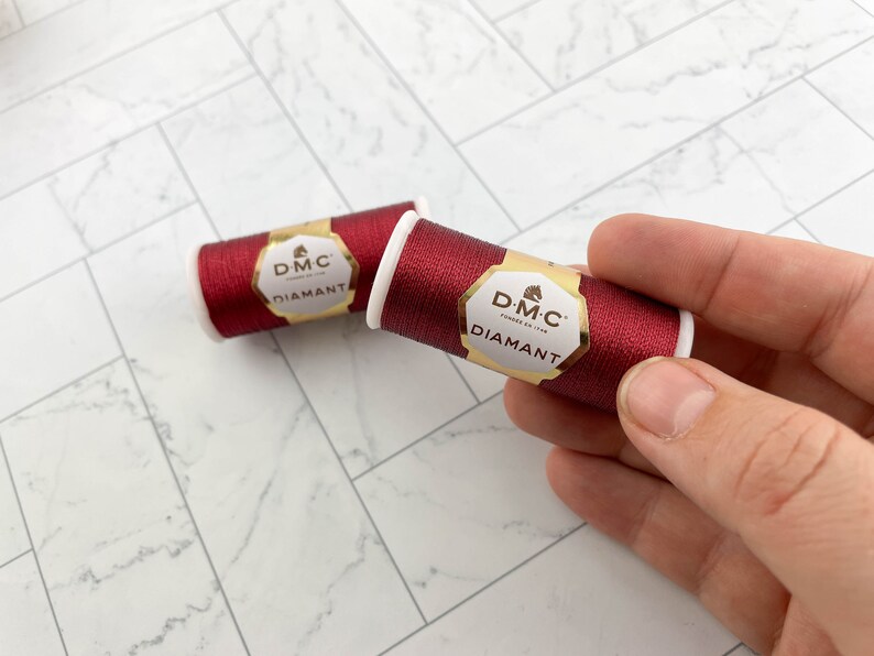 May include: Two spools of DMC Diamant thread in a deep red color. The spools have white cores and gold and white labels. The thread is held by a hand, with one spool in focus and the other slightly blurred in the background. The background is a white and gray patterned surface.