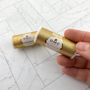 May include: Two spools of DMC Diamant metallic thread in gold. The spools are cylindrical with white end caps and gold labels. The labels feature the DMC logo and product information. The thread has a shimmering, metallic appearance.
