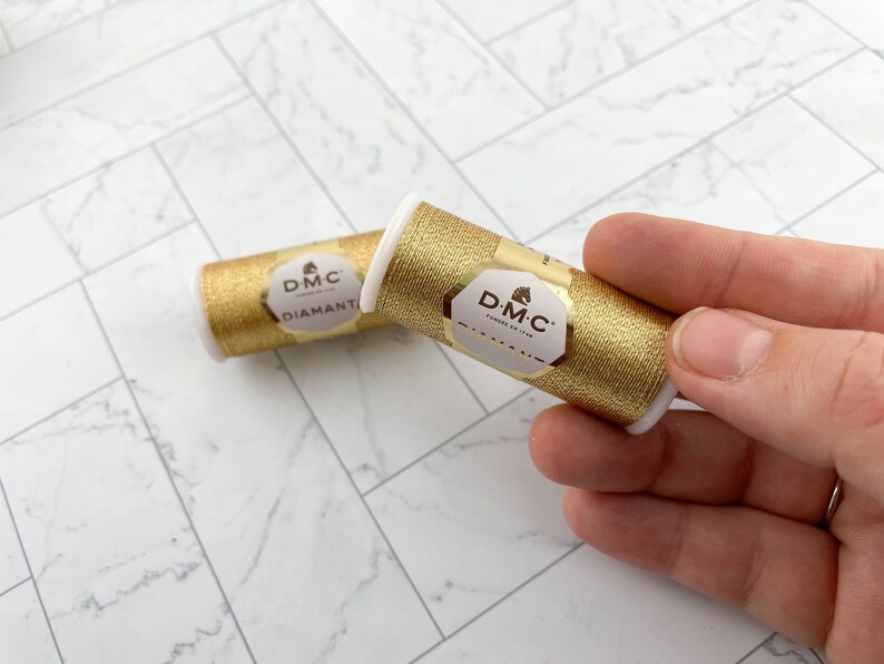 May include: Two spools of DMC Diamant metallic thread in gold. The spools are cylindrical with white end caps and gold labels. The labels feature the DMC logo and product information. The thread has a shimmering, metallic appearance.