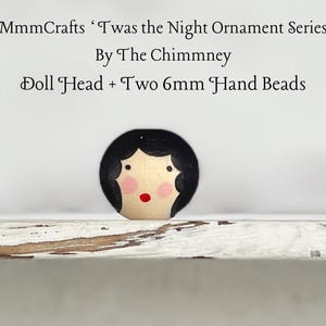 May include: A wooden doll head ornament with black hair, pink cheeks, and a red mouth. The ornament is part of a series called 'Twas the Night Ornament Series by The Chimmney. The ornament is accompanied by two 6mm hand beads.