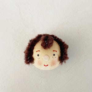 May include: A handmade doll head with a light beige face, rosy cheeks, and a small red smile. The doll has brown, fuzzy hair around the head. The eyes are small and black. This doll is a charming, handcrafted item.
