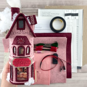 May include: A pink felt house kit with a white felt roof, a red felt door, and a window with a view of a bakery inside. The kit includes felt pieces, embroidery floss, and instructions for making a miniature house.