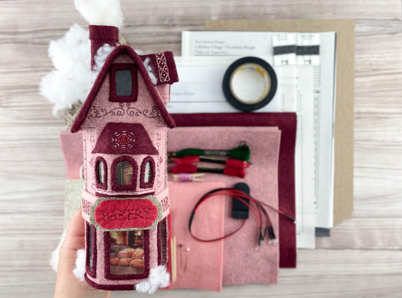 May include: A pink felt house kit with a white felt roof, a red felt door, and a window with a view of a bakery inside. The kit includes felt pieces, embroidery floss, and instructions for making a miniature house.