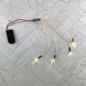 Peut inclure: Un ensemble de quatre lumières LED blanches de fée avec un bloc de piles noir. Les lumières sont reliées au bloc de piles par des fils fins et blancs.