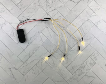 May include: A set of four white LED fairy lights with a black battery pack. The lights are connected to the battery pack by thin, white wires.