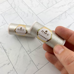 May include: Two spools of DMC Diamant metallic thread in silver. Each spool has a white plastic core and a gold and white label. The thread is held in a hand against a white and gray tiled surface.