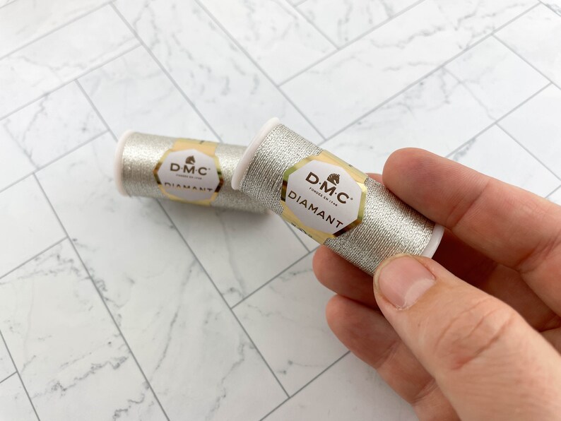 May include: Two spools of DMC Diamant metallic thread in silver. Each spool has a white plastic core and a gold and white label. The thread is held in a hand against a white and gray tiled surface.