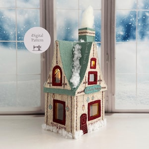 May include: A felt house decoration with a cream-colored exterior, a teal roof, and a chimney with white fiber smoke. The structure features red-trimmed windows, a red door, and embroidered ornaments. A sign reads Curiosity Shoppe.