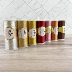 May include: A collection of DMC Diamant metallic embroidery thread spools in various colors, including silver, gold, red, pink, and brown. Each spool has a gold label with the DMC Diamant logo. The threads are arranged on a light wood surface.