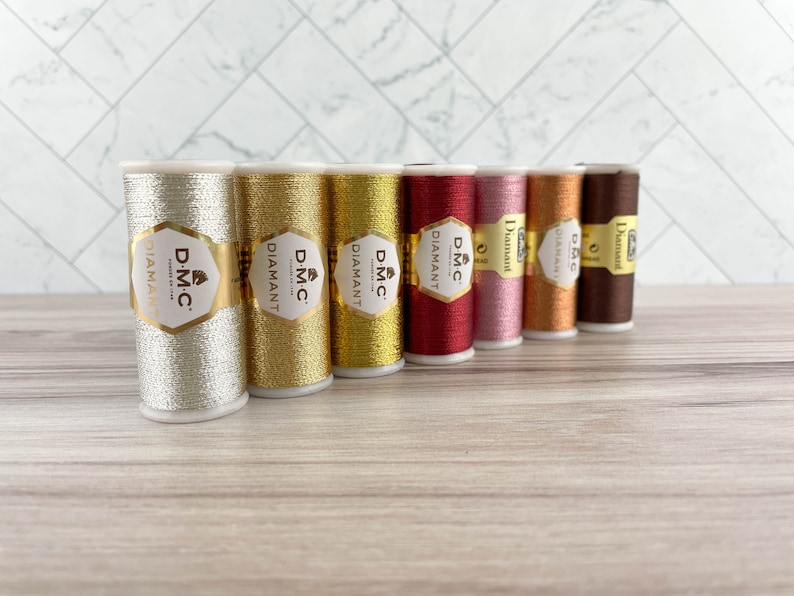 May include: A collection of DMC Diamant metallic embroidery thread spools in various colors, including silver, gold, red, pink, and brown. Each spool has a gold label with the DMC Diamant logo. The threads are arranged on a light wood surface.