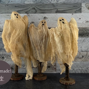 May include: Three handmade ghost decorations made from beige fabric with black eye details. Each ghost is mounted on a wooden stand. The background is a weathered wood panel. The image includes the text "Handmade!"