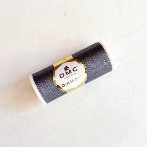 May include: A spool of DMC Diamant metallic thread in a dark gray color. The spool has white ends and a gold and white label with the DMC logo and the word "Diamant". The thread is designed for embroidery and other crafts.
