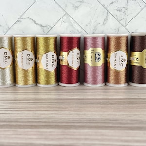May include: Seven spools of DMC Diamant metallic thread in shades of silver, gold, red, pink, copper, and brown. Each spool features a gold label with the DMC Diamant logo. The threads are displayed on a light wood surface.