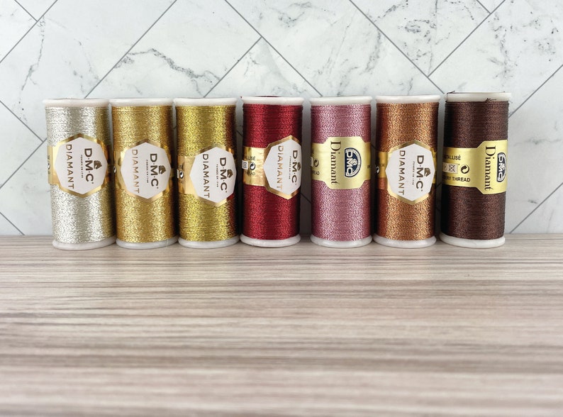 May include: Seven spools of DMC Diamant metallic thread in various colors, including silver, gold, red, pink, copper, and brown. Each spool has a gold label with the DMC Diamant logo. The threads are arranged on a light wood surface.