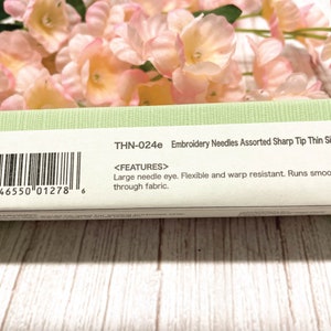 Puede incluir: Un paquete verde y blanco de agujas de bordar con el texto "THN-024e Embroidery Needles Assorted Sharp Tip Thin Sizes" y un c&oacute;digo de barras. El paquete tambi&eacute;n incluye el texto "<FEATURES> Large needle eye. Flexible and warp resistant. Runs smoothly through fabric."