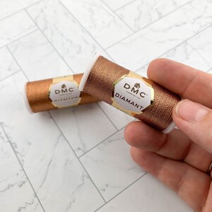May include: Two spools of DMC Diamant metallic embroidery thread in a copper color. The spools are cylindrical with white ends and a gold and cream label. The thread is held in a hand against a white and gray marble-patterned surface.