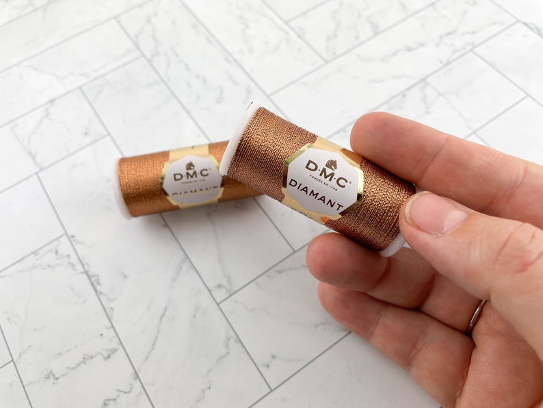 May include: Two spools of DMC Diamant metallic embroidery thread in a copper color. The spools are cylindrical with white ends and a gold and cream label. The thread is held in a hand against a white and gray marble-patterned surface.
