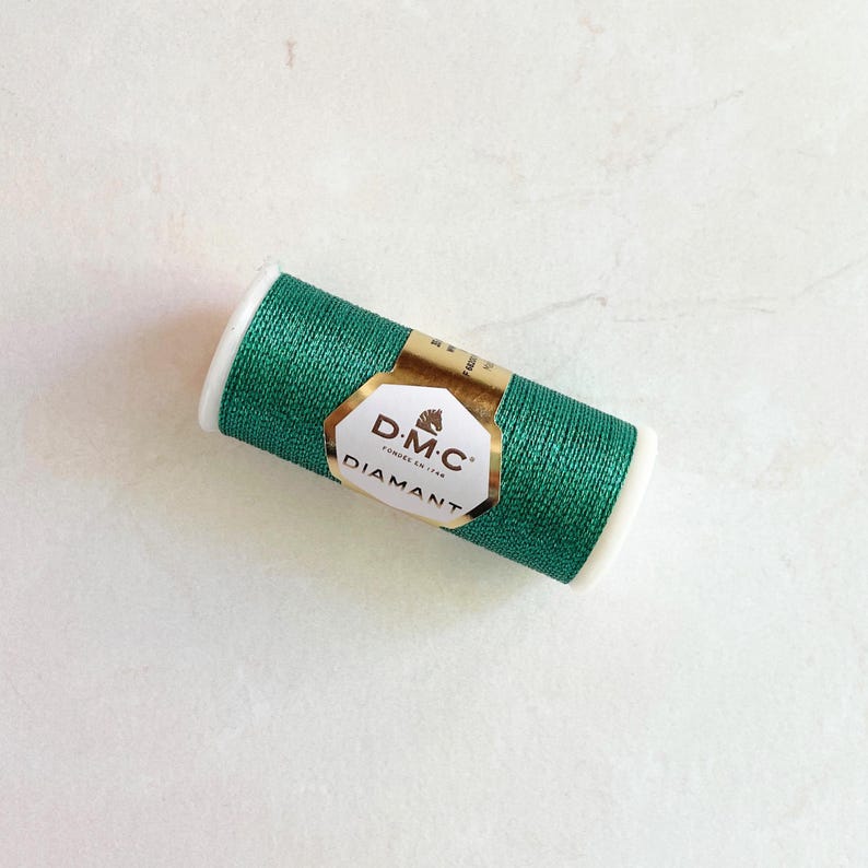 May include: A spool of emerald green DMC Diamant thread. The thread is wrapped around a white spool with a gold and white label. The label features the DMC Diamant logo.
