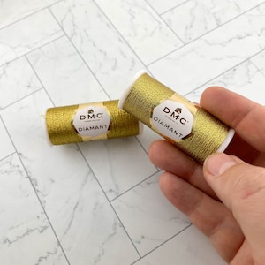 May include: Two spools of gold DMC Diamant thread. The spools are cylindrical with white ends and gold thread wrapped around them. Each spool has a label with the DMC Diamant logo. One spool is held by a hand.