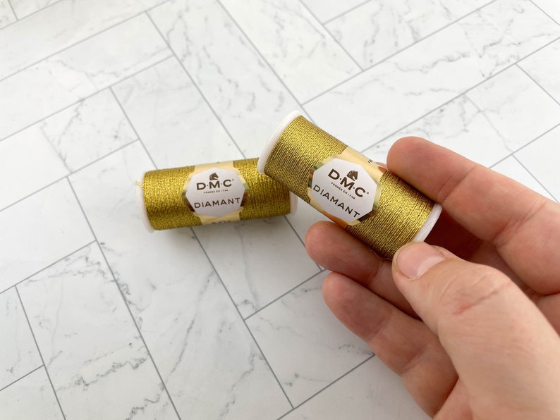 May include: Two spools of gold DMC Diamant thread. The spools are cylindrical with white ends and gold thread wrapped around them. Each spool has a label with the DMC Diamant logo. One spool is held by a hand.
